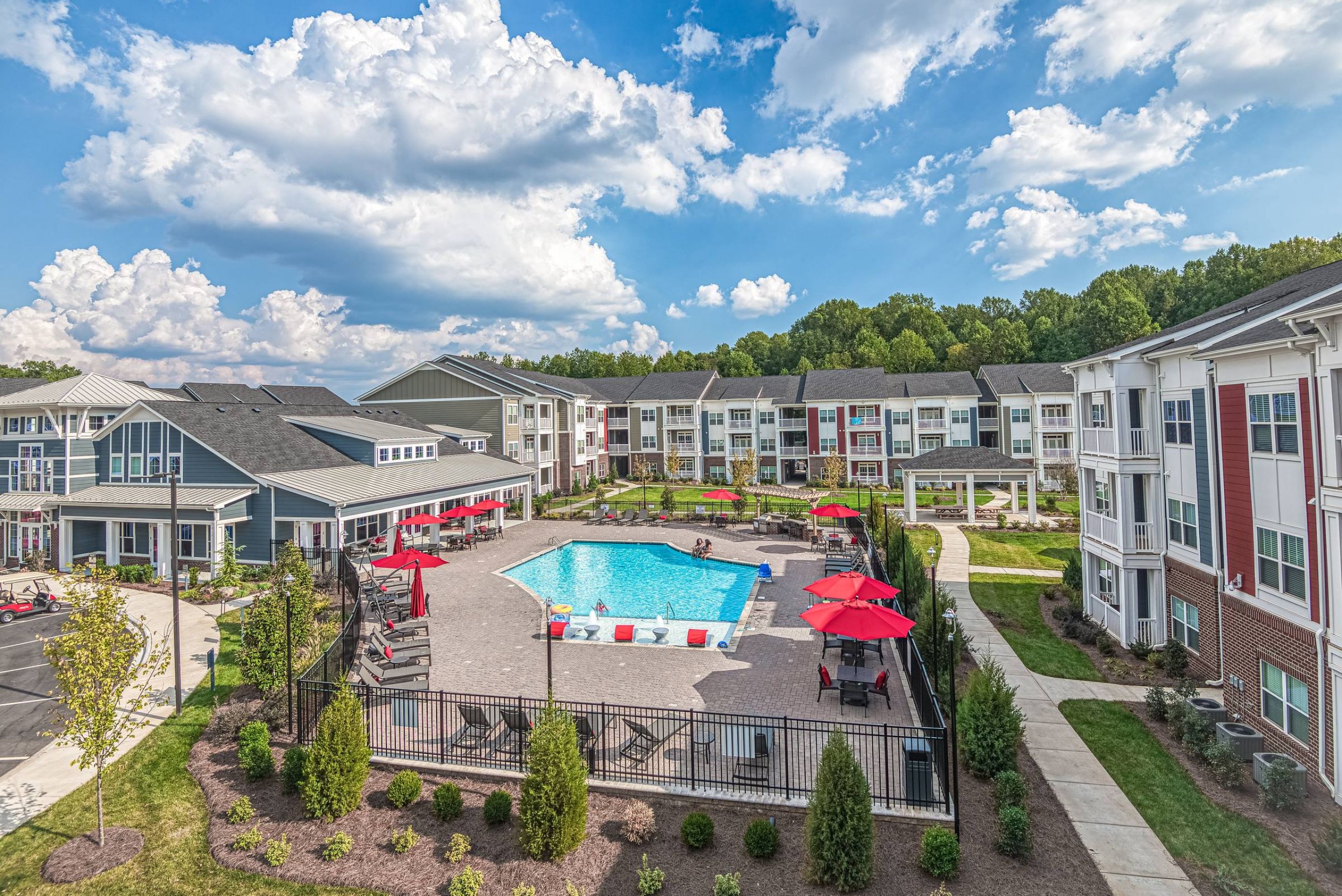 A modern apartment complex with a central outdoor pool, lounge chairs, and red umbrellas on a sunny day.