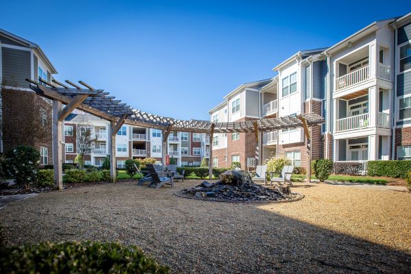 Courtyard with gravel, firepit, wooden pergola, chairs, and modern apartment buildings under a clear blue sky.