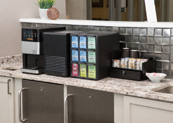Coffee and tea station with a hot beverage machine, assorted tea packets, and creamers on a granite counter.