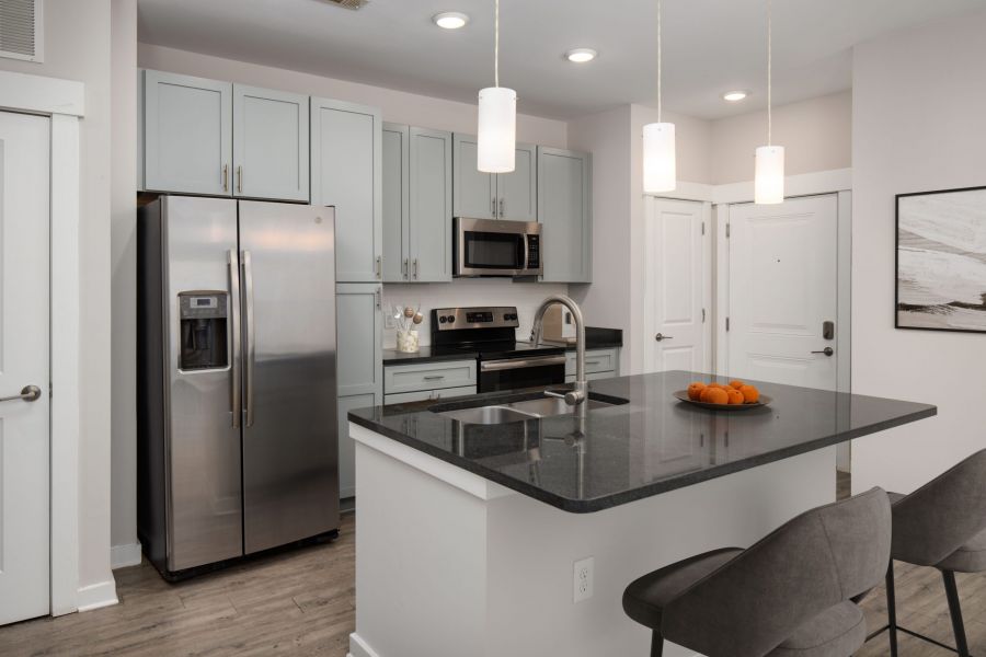 Modern kitchen with gray cabinets, stainless steel appliances, and an island with two chairs and a bowl of oranges.