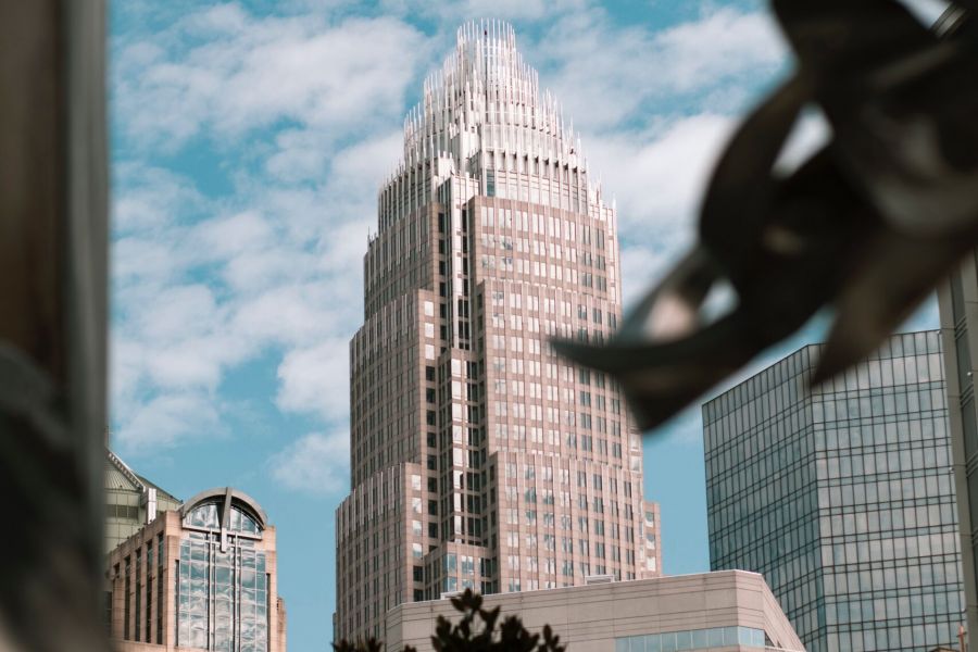 Tall skyscraper with a tiered, pointed top against a blue sky with clouds, framed by nearby buildings and leaves.