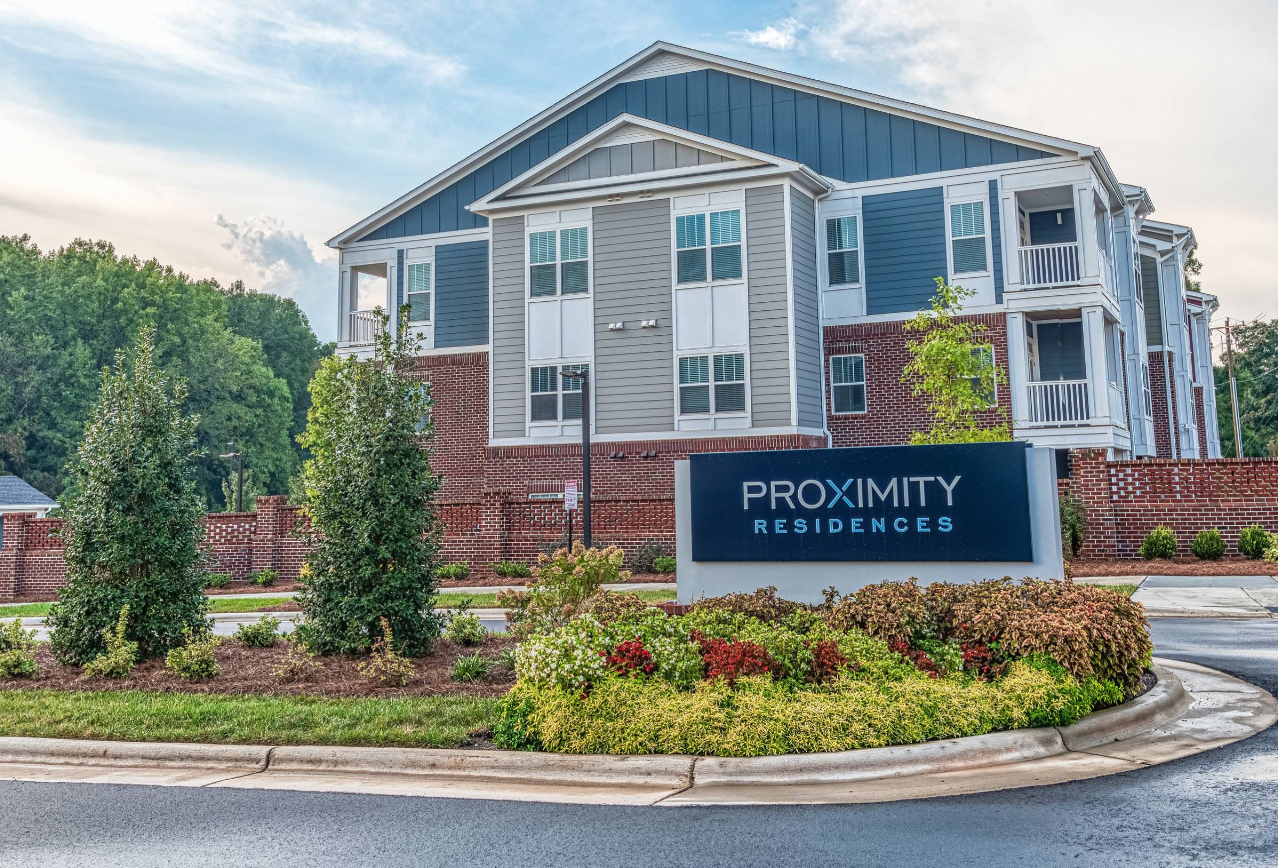Modern apartment building with a sign reading "Proximity Residences," surrounded by landscaped greenery.