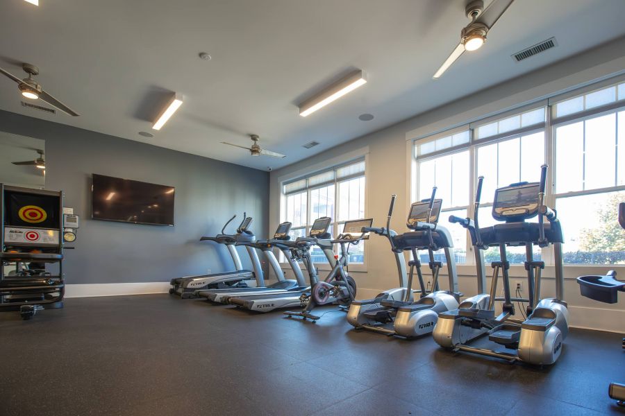 Modern gym with treadmills, elliptical machines, a TV, large windows, and bright overhead lighting.