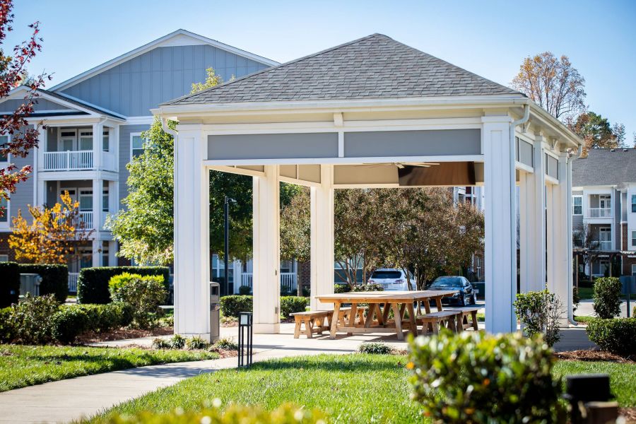 A wooden gazebo with picnic tables in a sunny residential complex, surrounded by greenery and parked cars.