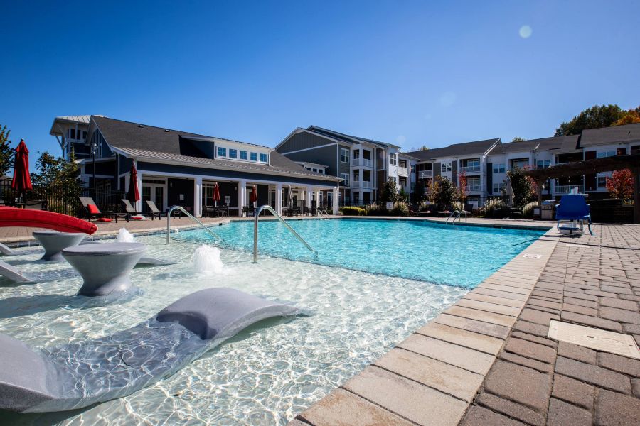 Outdoor swimming pool with lounge chairs, fountains, and modern apartment buildings in the background under clear sky.